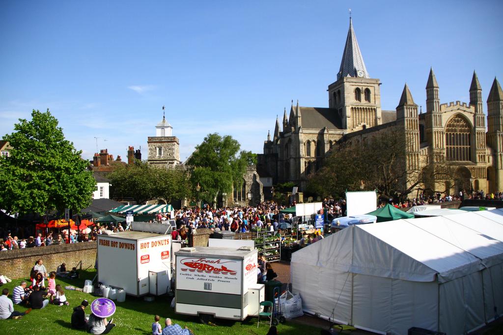 Historische Gebäude mit Bäumen davor, ein Turm im Hintergrund, Menschen auf einem Weg mit Geschäften daneben und andere auf der Wiese in der Nähe sitzend.