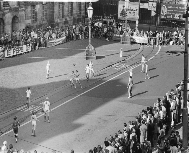 Schwarzes Foto eines Streetsoccer-Spiels mit spielenden Fußballern und Zuschauern von der Seitenlinie aus, umgeben von Laternenmasten, Gebäuden und Bannern.