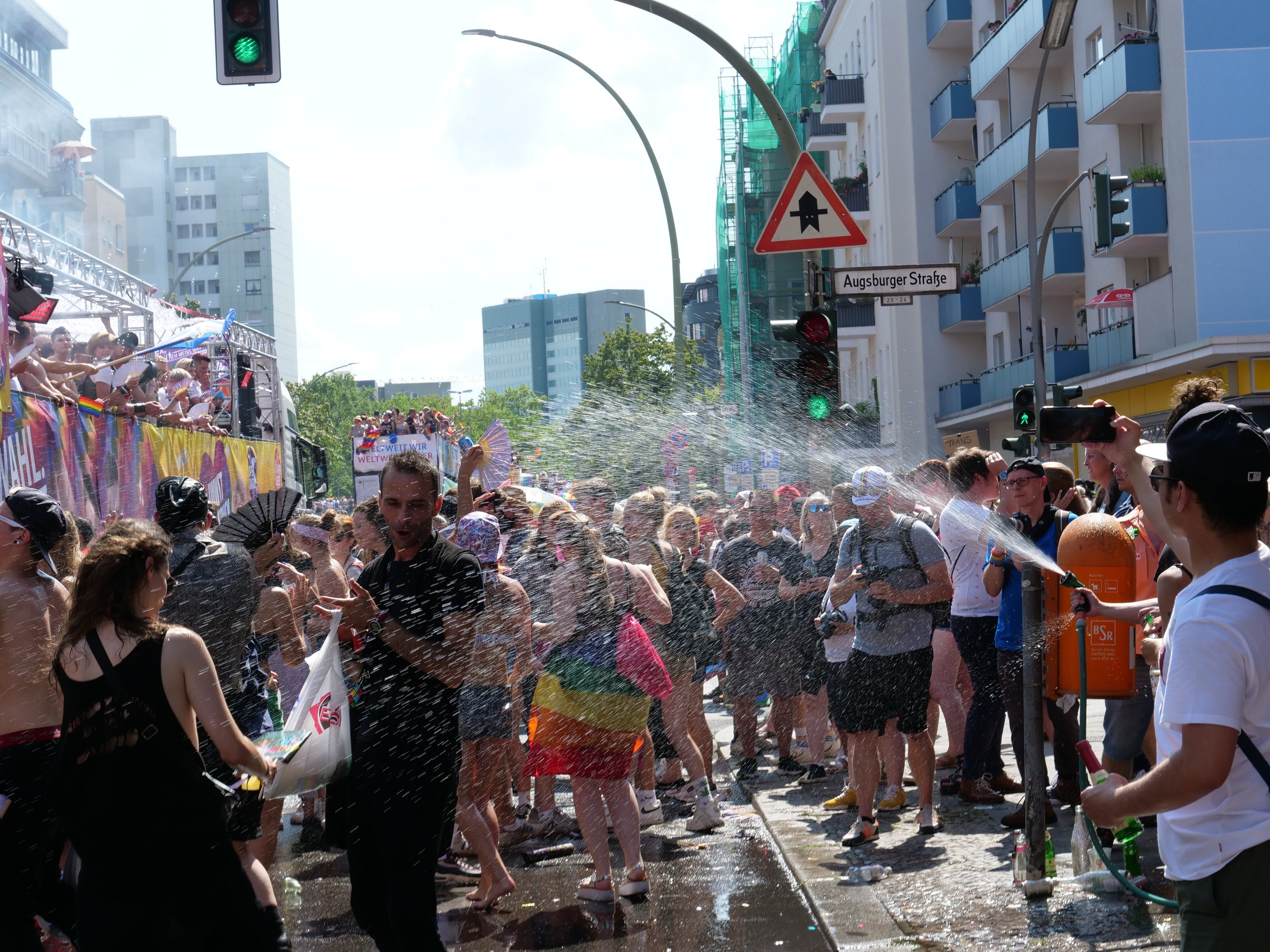 Gruppe von Menschen auf einer Christopher Street Day Parade, die sich gegenseitig mit Wasser bespritzen und Gegenstände halten, mit einem Banner links und Gebäuden, Bäumen und Straßenelementen im Hintergrund.