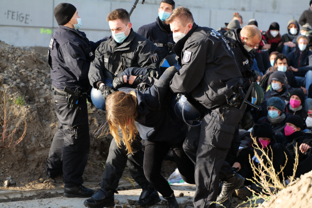 Eine Gruppe maskierter Personen in Mützen, einige bewaffnet, umgibt eine Frau, die von der Polizei eskortiert wird, mit Pflanzen, Steinen und einer Wand mit Protesttext im Hintergrund.