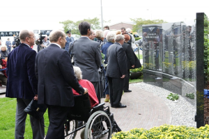 Ein Mann im Anzug sitzt in einem Rollstuhl in der Mitte einer Gruppe von Menschen, die um eine Gedenktafel mit Text stehen, umgeben von Kameras und Blumen, mit Bäumen, Gebäuden und einem klaren blauen Himmel im Hintergrund.