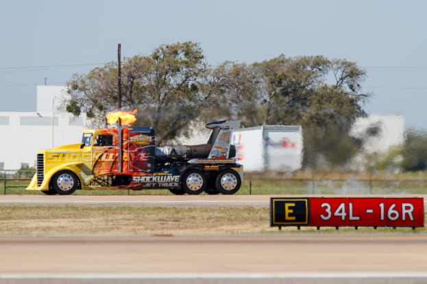 Ein Lkw mit Flammen am Hückenden auf einer Straße mit Gras, einem Schild, einem Zaun, Bäumen, Mäständen, Drähten, einem Gebäude und Himmel im Hintergrund.