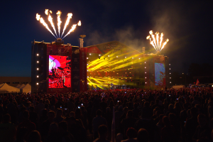 Eine große Menschenmenge auf einem Musikfestival mit einer Bühne, auf der Künstler performen, helles Licht und Banner, Zelte links und Feuerwerk am dunklen Himmel.
