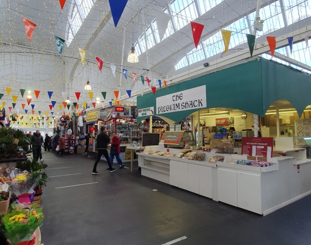 Innenmarkt-Szene mit Menschen, Blumensträußen links, Essenswaren auf Tischen rechts, einem Stand mit Namensschild und dekorativen Fahnen an der Decke.