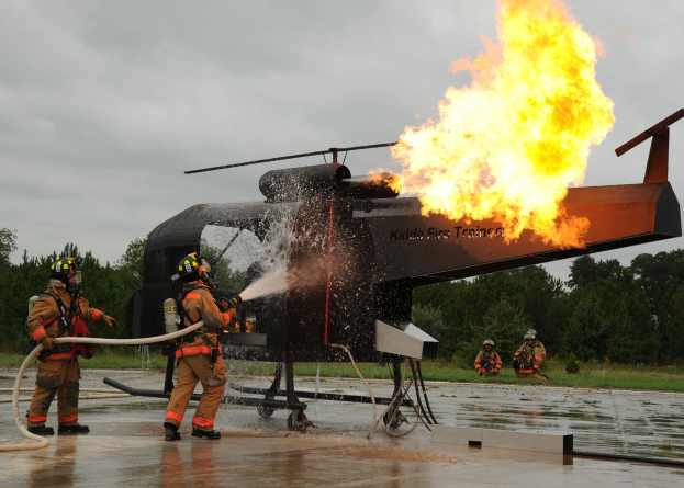 Feuerwehrleute spritzen Wasser auf einen brennenden Hubschrauber, während im Hintergrund Bäume und ein klarer Himmel zu sehen sind.