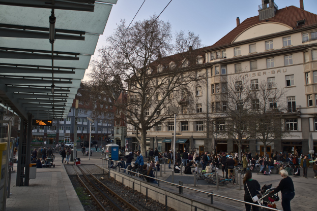 Eine belebte Straßenkreuzung mit einem Bahnhof auf der linken Seite, umgeben von Gebäuden, Bäumen, Laternenmasten und Textanzeigen, wo Menschen gehen, auf Bänken sitzen oder stehen.