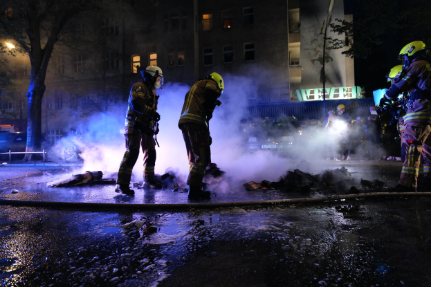 Feuerwehrleute in Helmen stehen nachts auf einer Straße mit Rauch aus einer Leitung auf der Straße, halten Gegenstände, während Gebäude, Bäume und Fahrzeuge im Hintergrund zu sehen sind mit einer Tafel rechts.