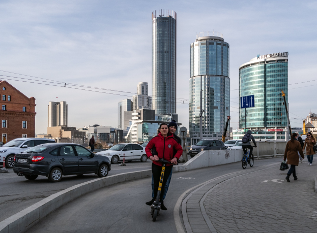 Ein Mann in einer roten Jacke und schwarzen Hosen fährt mit einem Elektroroller die Straße mit hohen Gebäuden hinunter, umgeben von Passanten und Radfahrern unter einem teilweise bewölktem Himmel.
