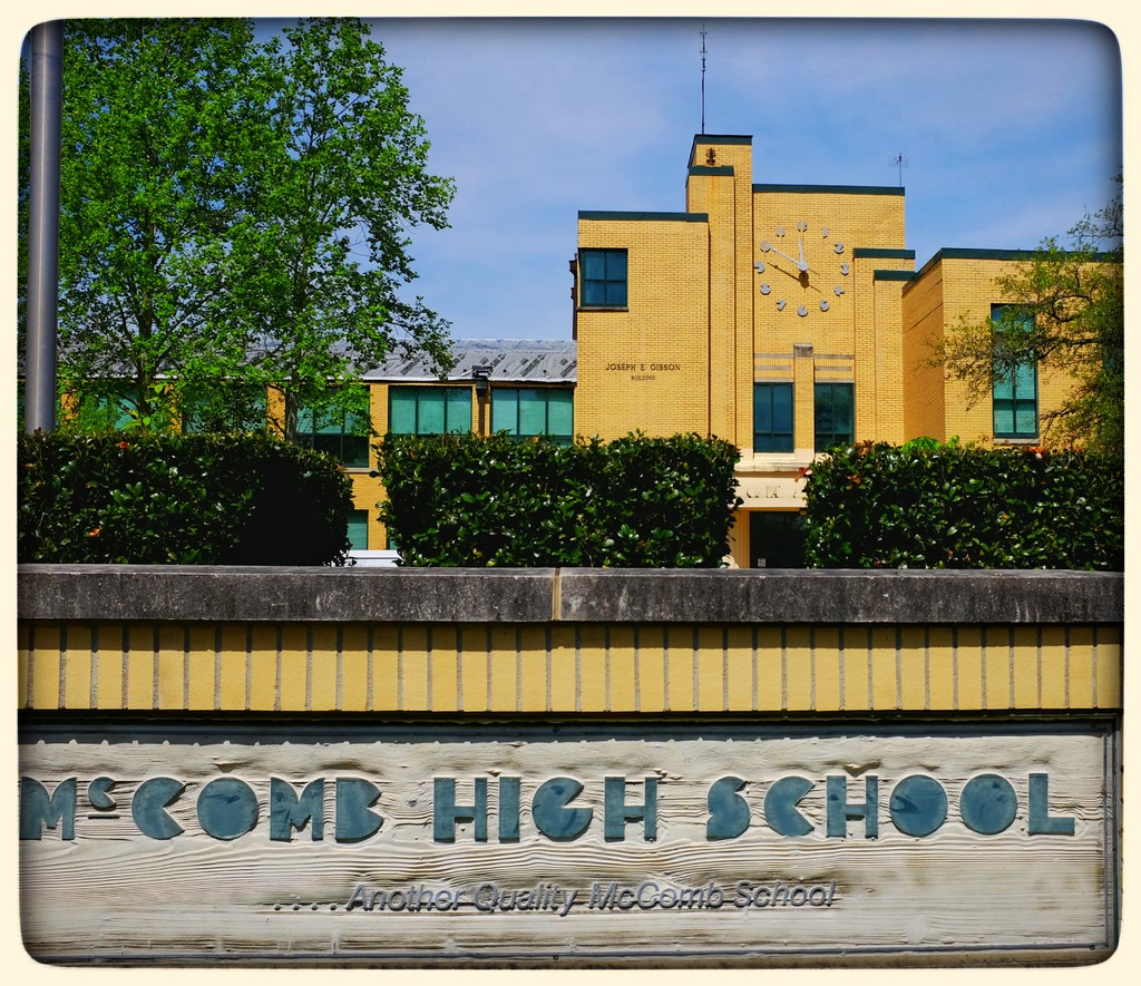 Ein Plakat mit dem Text "MCCOMB HIGH SCHOOL" auf einer Wand, umgeben von Pflanzen, Bäumen, einem Pfahl, einem Gebäude und einem bewölkten Himmel.