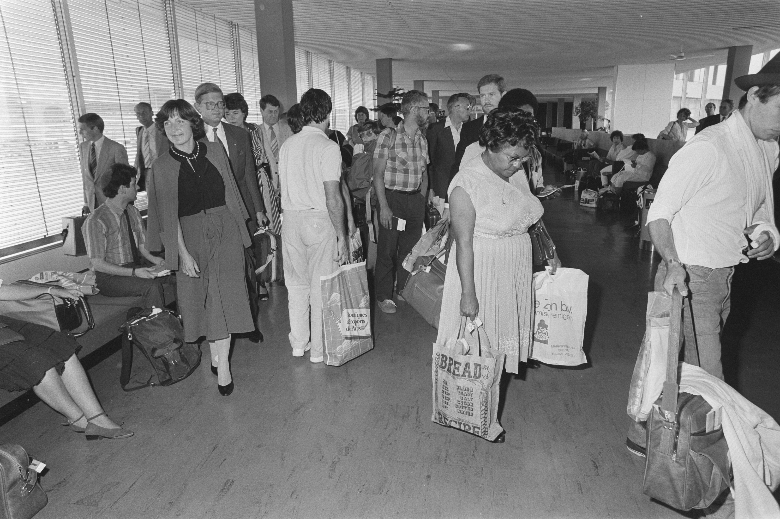Menschen gehen und sitzen in einem schwarz-weißen Flughafen-Terminal mit Jalousien auf der linken Seite, ein Mann und eine Frau stehen in der Mitte und halten jeweils eine Tasche.