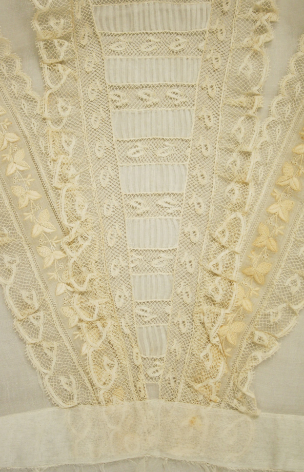 Nahaufnahme eines weißen Kleides mit zarten, detaillierten Spitzenverzierungen auf einem leichten, luftigen Stoff.