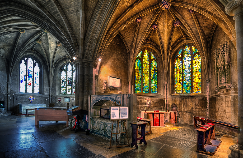 Ein Raum mit Bänken, Tischen, einer Tafel, Fenstern, einer Wand, Kerzen, einem Kamin und Lichtern in der Mitte, bearbeitet.