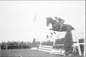 Schwarzes und weißes Foto eines Pferdes und Reiters, die über ein Hindernis bei den Royal Ascot Horse Trials 1953 springen, mit Zuschauern und einer Fahne im Hintergrund.