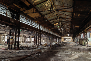 Ein verlassener Fabrikinnenraum mit verstreuter Trämmerei, kaputten Fenstern, Metallstäben und Deckenlampen unter einem beschädigten Dach.